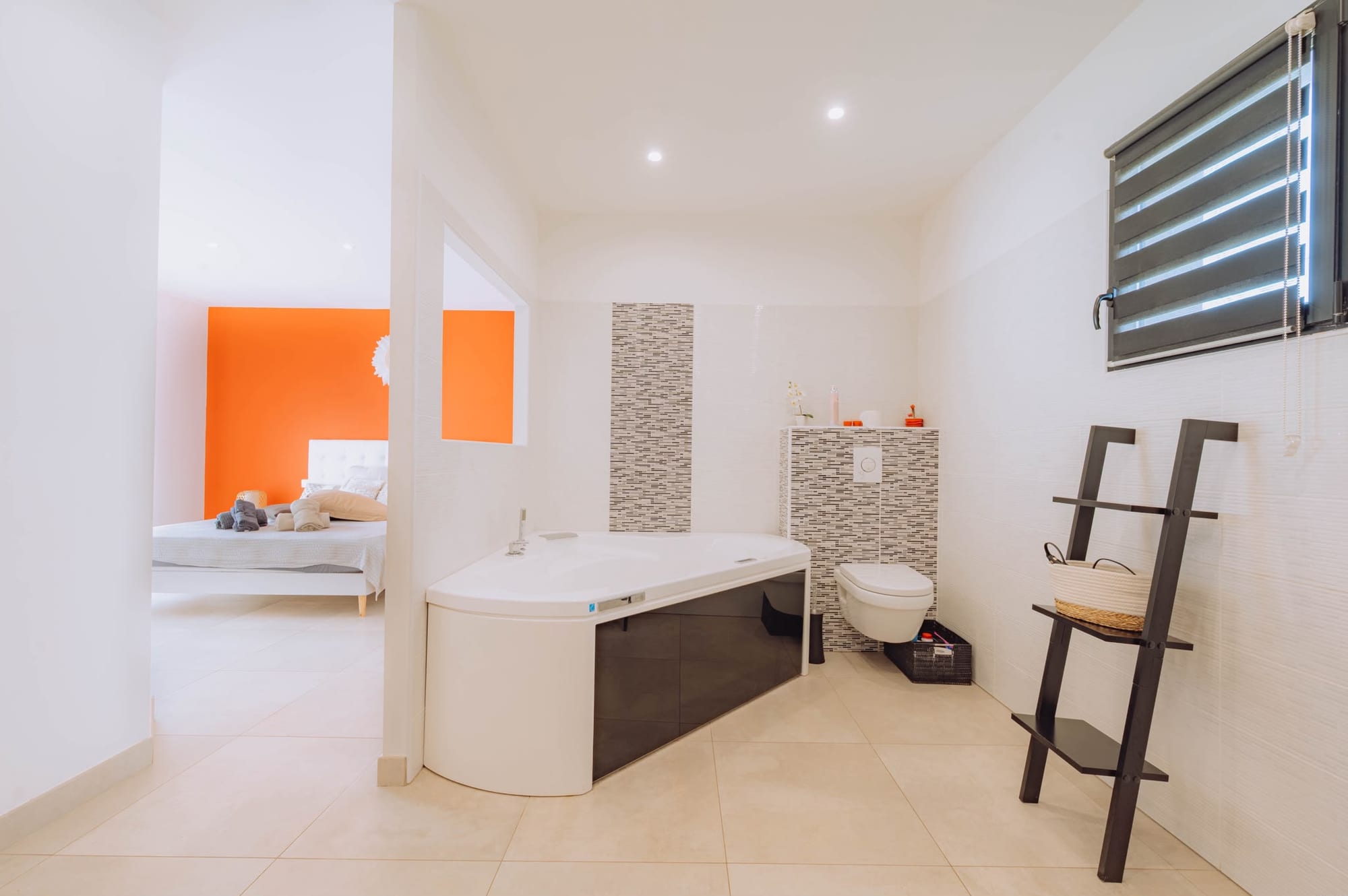 Salle de bain avec baignoire d'angle, WC suspendu et frise en mosaïque, ouverte sur la chambre